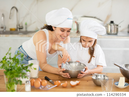 Mom and girl in white chief hats are talking in kitchen and whisking ingredients for pastry in bowl 114290334