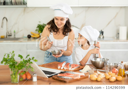 While cooking salmon fillet fish dish, woman and little daughter in hats look at laptop While cooking salmon fillet fish dish, woman and little daughter in hats look at laptop 114290399
