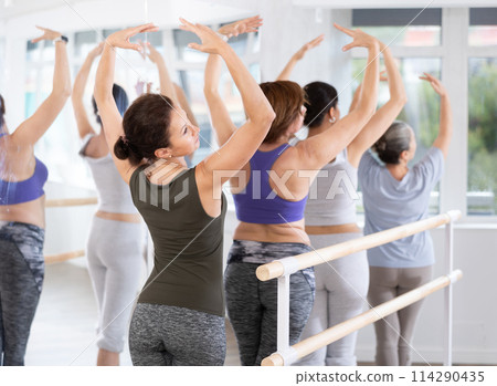 Ordinary women, not professionals, stand in fifth position near ballet barre during group training in dance studio 114290435