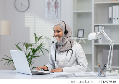 A professional and cheerful female doctor in a hijab provides online consultations from her well-organized office, using a laptop and headset. 114297646
