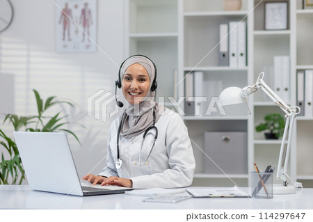 A professional female doctor wearing a hijab, with a stethoscope, sits smiling at her desk in a bright, modern clinic office, using a laptop and wearing a headset. A professional female doctor wearing a hijab, with a stethoscope, sits smiling at her desk in a bright, modern clinic office, using a laptop and wearing a headset. 114297647
