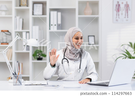 Cheerful Muslim female doctor in hijab waving hello during an online consultation from her well-organized office. Modern healthcare and telemedicine concept. 114297663