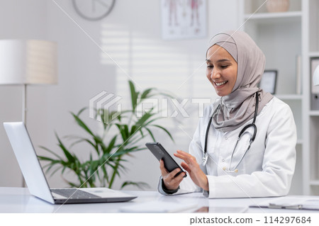 Cheerful Muslim female doctor in hijab uses smartphone while sitting at her desk. Modern, well-lit clinic office setting celebrates diversity and technology in healthcare. Cheerful Muslim female doctor in hijab uses smartphone while sitting at her desk. Modern, well-lit clinic office setting celebrates diversity and technology in healthcare. 114297684