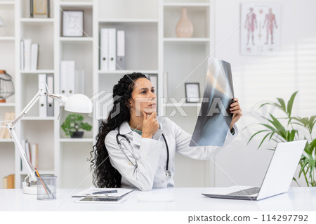 Focused Hispanic female doctor examining an X-ray film while contemplating diagnostics in a well-equipped modern medical office. Focused Hispanic female doctor examining an X-ray film while contemplating diagnostics in a well-equipped modern medical office. 114297792