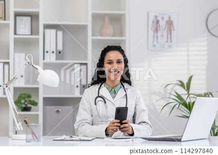A professional Hispanic female doctor smiling at camera, holding a smartphone in a modern medical office. She exudes friendliness and competence. A professional Hispanic female doctor smiling at camera, holding a smartphone in a modern medical office. She exudes friendliness and competence. 114297840