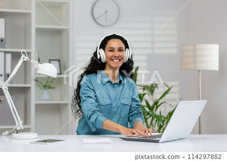 A cheerful Hispanic woman wearing headphones works at her desk in a bright modern office, interacting with her laptop, embodying professionalism and happiness. 114297882