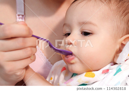 Portrait of little baby boy eating food. Baby with a spoon in feeding chair. Cute baby eating first meal Portrait of little baby boy eating food. Baby with a spoon in feeding chair. Cute baby eating first meal 114302818