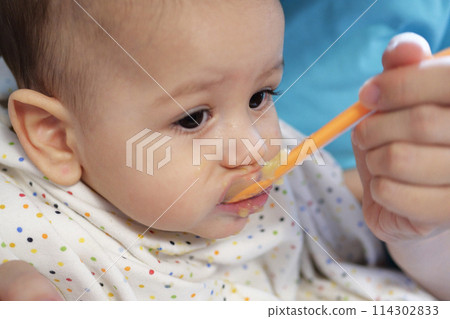 Portrait of little baby boy eating food. Baby with a spoon in feeding chair. Cute baby eating first meal 114302833