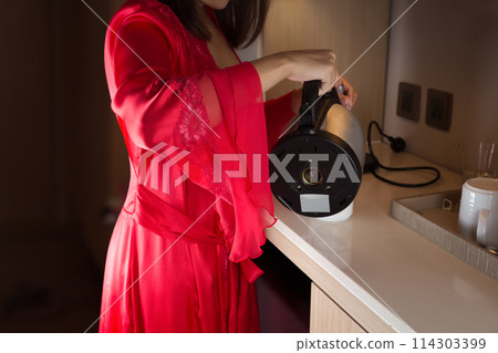 A woman in a nightgown is standing, pouring hot water into a white cup. 114303399