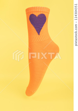 Orange cotton sock on yellow background. volumetric sock with purple heart. advertising, logo, branding. 114304351
