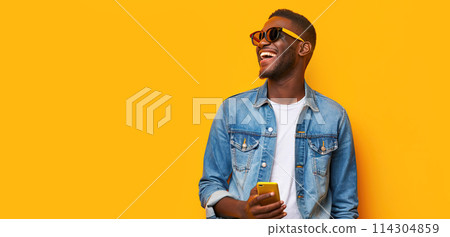 Happy African millennial man laughing with mobile phone on yellow studio backdrop. Joyful black man wearing jeans jacket, sunglasses smiling looking aside. Great new mobile App. People and gadgets 114304859