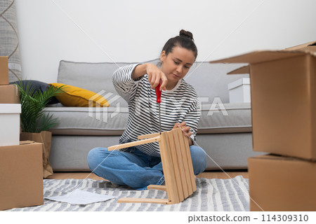 Young brunette woman installs furniture using construction tools sorting out carton boxes of things for cozy interior in new living space in purchased apartment 114309310