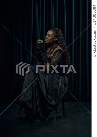 African-American woman in vintage outfit, looks as medieval person holds chocolate donut against dark curtain backdrop. 114309394