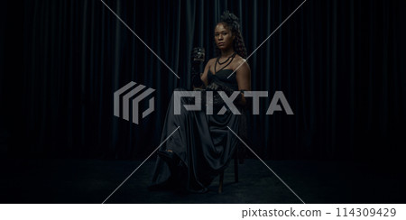 African-American woman, dressed old-fashion dress, looks medieval person, sits confidently on chair holding glass of brandy against dark curtain backdrop. 114309429