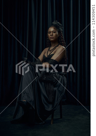 African-American woman, dressed old-fashion dress, looks as princess, sits confidently on chair against dark curtain backdrop. 114309451