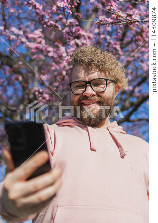 Spring day. Bearded man in pink shirt talking by phone. Spring pink sakura blossom. Handsome young man with smartphone. Fashionable man in trendy glasses. Bearded stylish man. Copy space 114309874