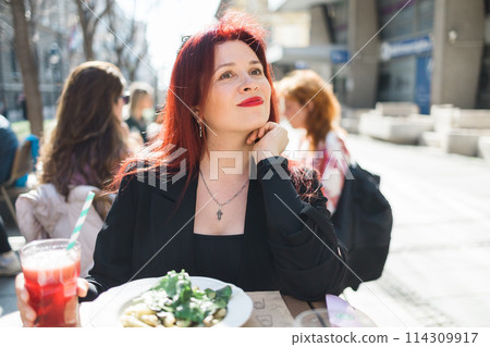 Beautiful happy millennial woman with long red hair enjoying italian pasta in a street cafe 114309917