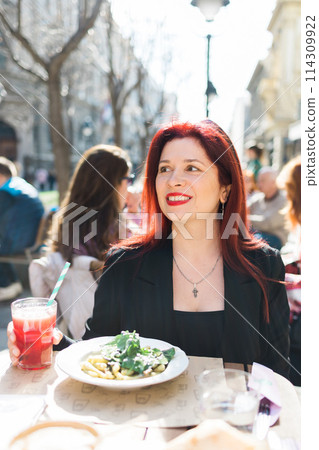 Beautiful happy millennial woman with long red hair enjoying italian pasta in a street cafe 114309922