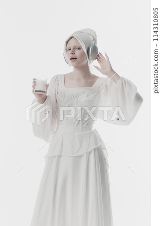 Young lady, beautiful woman wearing old fashion clothes listening music in headphones and drinks milk against white studio background. Young lady, beautiful woman wearing old fashion clothes listening music in headphones and drinks milk against white studio background. 114310805