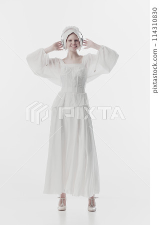 Young lady, in guise of simple peasant woman wearing old fashion clothes listening music in headphones against white studio background. Young lady, in guise of simple peasant woman wearing old fashion clothes listening music in headphones against white studio background. 114310830