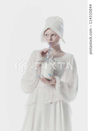 Beautiful young woman dressed as medieval person, maid in white historic dress drinks milk against white studio background. Beautiful young woman dressed as medieval person, maid in white historic dress drinks milk against white studio background. 114310844