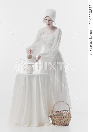 Young woman dressed as medieval person, maid in white dress poring milk into jug against white studio background. 114310855