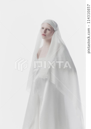 Young, calm woman shrouded in gossamer white fabric, creating ghostly yet angelic appearance against white studio background. 114310857