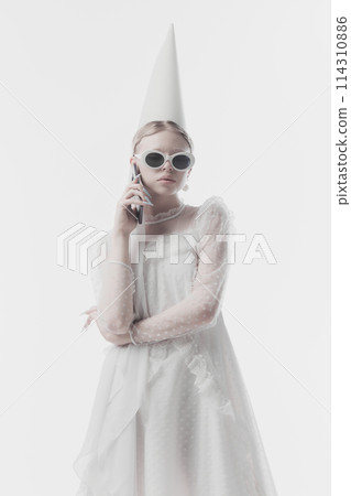 Young woman in white dress with conical hat, and modern sunglasses, looks as medieval person, talking on phone against white studio background. 114310886