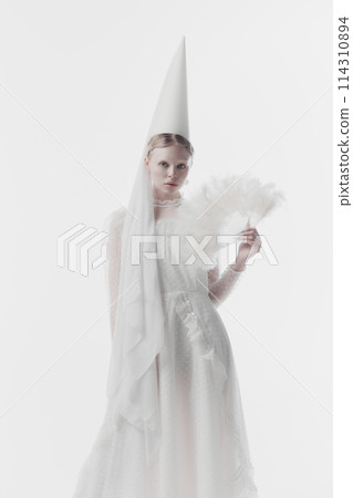 Portrait of young woman in white costume with conical hat, looks as medieval person, holding feathery fan against white studio background. Portrait of young woman in white costume with conical hat, looks as medieval person, holding feathery fan against white studio background. 114310894