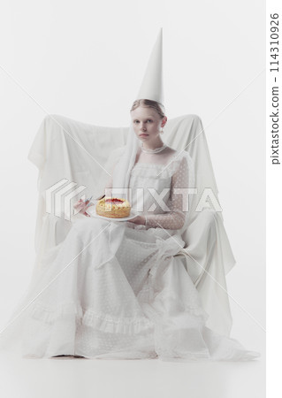Woman in elaborate white gown and pointed hat, looks as medieval person, sits, holding sweet cake against white studio background. Woman in elaborate white gown and pointed hat, looks as medieval person, sits, holding sweet cake against white studio background. 114310926