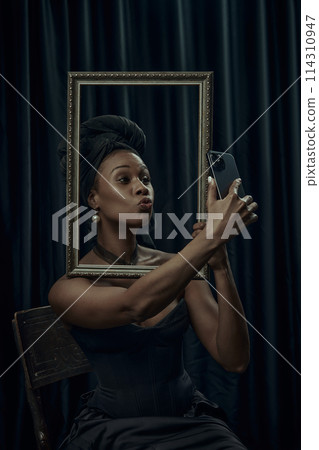 African-American woman sitting posing behind ornate, vintage frame and taking selfie using modern smartphone against dark curtain backdrop. African-American woman sitting posing behind ornate, vintage frame and taking selfie using modern smartphone against dark curtain backdrop. 114310947