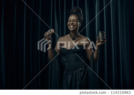 Smiling African-American woman, dressed in vintage black dress holds sweet donut and cup of coffee against dark curtain backdrop. Smiling African-American woman, dressed in vintage black dress holds sweet donut and cup of coffee against dark curtain backdrop. 114310963