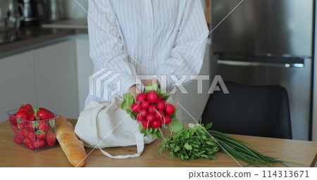 Woman put a bag of groceries on the kitchen table. Women hands put greens and vegetables on the table. Woman put a bag of groceries on the kitchen table. Women hands put greens and vegetables on the table. 114313671
