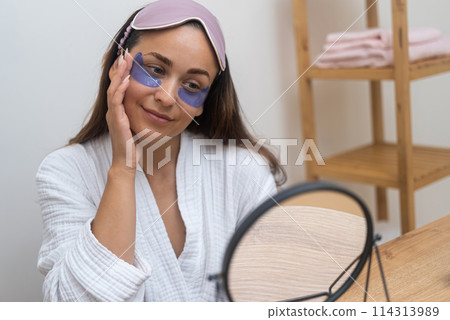 Radiant Revival: A woman starts her day with a mirror's gaze, using under-eye patches to address puffiness, wrinkles, and dark circles. Radiant Revival: A woman starts her day with a mirror's gaze, using under-eye patches to address puffiness, wrinkles, and dark circles. 114313989