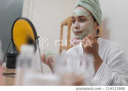 Self-Love Ritual: She, in a bathrobe, applies a clay mask with a brush, enjoying her organic facial treatment and the mirror's reflection. Self-Love Ritual: She, in a bathrobe, applies a clay mask with a brush, enjoying her organic facial treatment and the mirror's reflection. 114313990