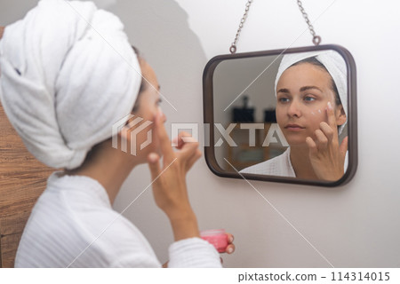 Home skincare: A woman, her reflection in the mirror, diligently applies face moisturizer, elevating her routine. Home skincare: A woman, her reflection in the mirror, diligently applies face moisturizer, elevating her routine. 114314015