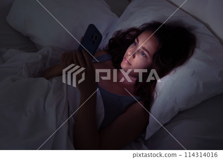 In the silent night, a woman lies in bed, drawn to her phone's glow, a scene illustrating the grip of insomnia and social media addiction.  114314016