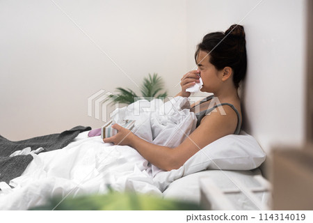 A sick woman uses tissues to wipe her nose while holding medical drops, seeking relief in her bedridden state 114314019