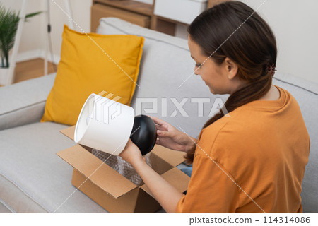 Woman takes out small table lamp from cardboard parcel with home delivery online purchase from furniture store and decorative element with lighting function Woman takes out small table lamp from cardboard parcel with home delivery online purchase from furniture store and decorative element with lighting function 114314086