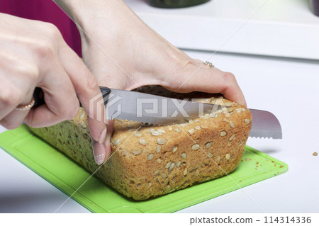 Baking bread at home. A loaf of freshly baked wholegrain bread with sunflower seeds lies on a cutting board. A woman cuts it with a knife. Baking bread at home. A loaf of freshly baked wholegrain bread with sunflower seeds lies on a cutting board. A woman cuts it with a knife. 114314336