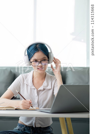 Smiling girl student wear headphone study online with teacher, happy young asian woman learn lecture watch webinar write notes look at laptop sit in home, distant education 114315668