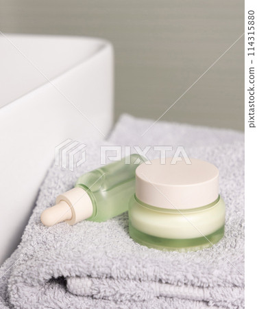 Light green dropper bottle and cream jar on light grey bath towel near basin closeup Light green dropper bottle and cream jar on light grey bath towel near basin closeup 114315880