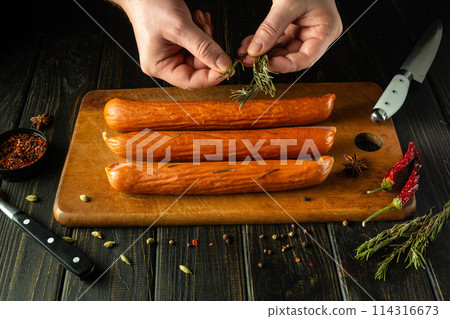 The cook adds aromatic rosemary to the smoked sausages with his hands. Low key concept of grilling for lunch. Place for advertising The cook adds aromatic rosemary to the smoked sausages with his hands. Low key concept of grilling for lunch. Place for advertising 114316673