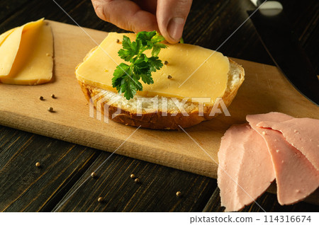 Cooking sandwiches with sausage and cheese on the kitchen table by the hands of a cook. Low key concept of preparing quick food for snacking Cooking sandwiches with sausage and cheese on the kitchen table by the hands of a cook. Low key concept of preparing quick food for snacking 114316674