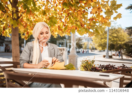 A blonde woman sits at a table with a cup of coffee and a leaf on it. The scene is set in a city with a tree in the background. 114316948
