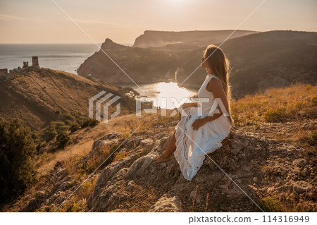 A woman in a white dress sits on a rock overlooking a body of water. The scene is serene and peaceful, with the woman enjoying the view and the calmness of the surroundings. A woman in a white dress sits on a rock overlooking a body of water. The scene is serene and peaceful, with the woman enjoying the view and the calmness of the surroundings. 114316949