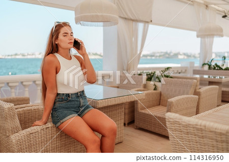 Cafe woman sitting at a table with a cell phone in her hand. She is looking at the screen and she is focused on her phone. The scene takes place in a restaurant or cafe, with multiple chairs. Cafe woman sitting at a table with a cell phone in her hand. She is looking at the screen and she is focused on her phone. The scene takes place in a restaurant or cafe, with multiple chairs. 114316950