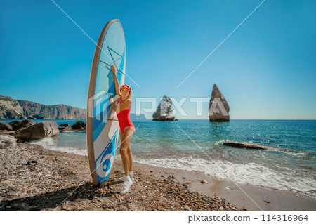 Woman surfer during summer travel to beach vacation. A middle-aged woman in a red swimsuit and a cap with sap on the background of the sea and rocks in the water. extreme sport. Travel, weekend Woman surfer during summer travel to beach vacation. A middle-aged woman in a red swimsuit and a cap with sap on the background of the sea and rocks in the water. extreme sport. Travel, weekend 114316964