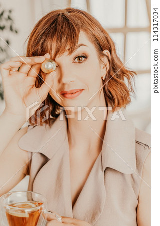 Woman drinks tea close-up. Portrait of a brunette in a beige dress with a transparent mug and a candy in her hands. Woman drinks tea close-up. Portrait of a brunette in a beige dress with a transparent mug and a candy in her hands. 114317034
