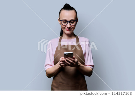 Portrait of 30s woman in apron holding smartphone in hands on grey background 114317168